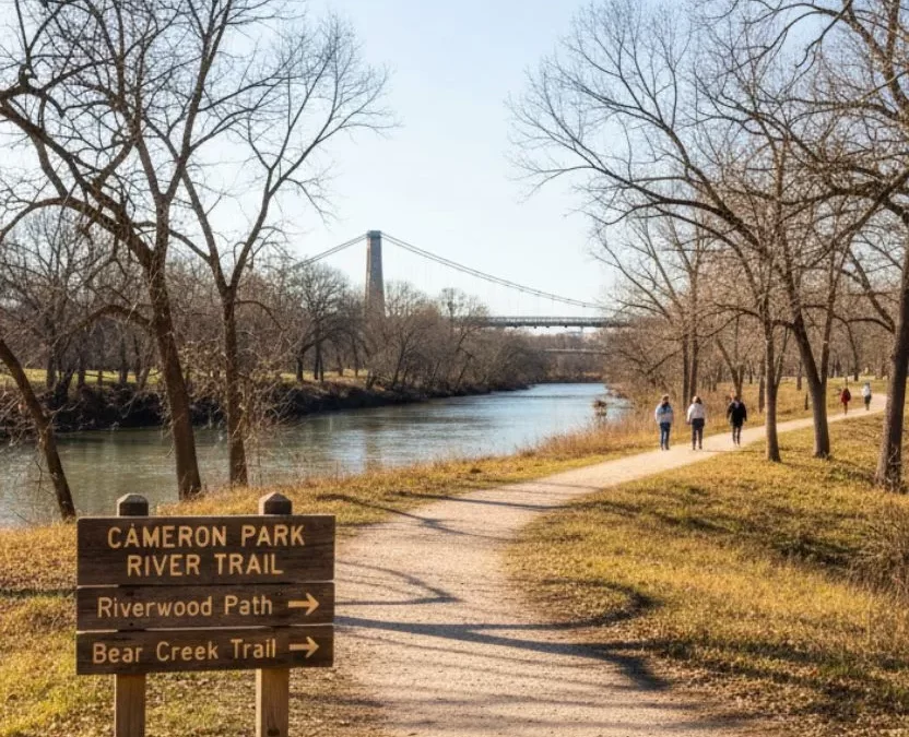 A family walks on a river trail in a Waco park during winter, with the historic Suspension Bridge and a partially frozen river visible in the background.