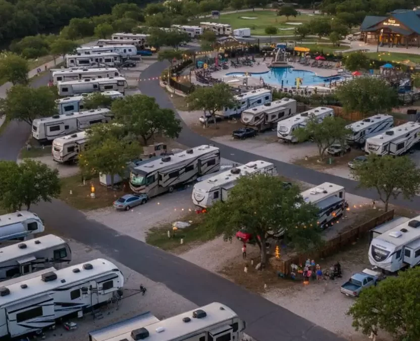 Aerial sunset view of Camp Fimfo Waco RV park featuring rows of modern RVs and fifth wheels parked on concrete pads along the Bosque River with the Waco city skyline and a bridge in the background.