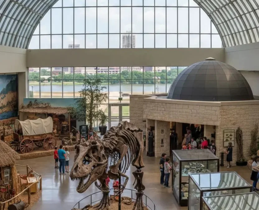 A wide-angle indoor view of a natural history museum in Waco, Texas, featuring a large dinosaur skeleton, historical dioramas, and a glass-domed ceiling overlooking the city.