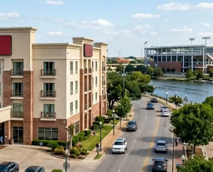 An aerial view of affordable hotels in Waco Texas near Brazos River, featuring a modern brick hotel building alongside a waterfront promenade and a stadium in the background.
