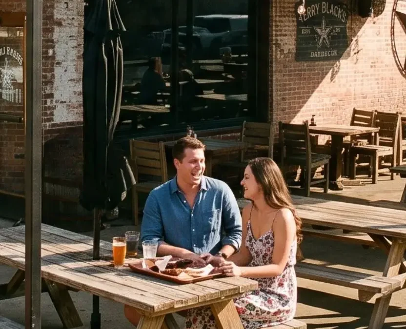 A couple sits at an outdoor picnic table on the patio of a brick barbecue restaurant in downtown Waco, Texas, laughing over a tray of food and beers under a string of patio lights.